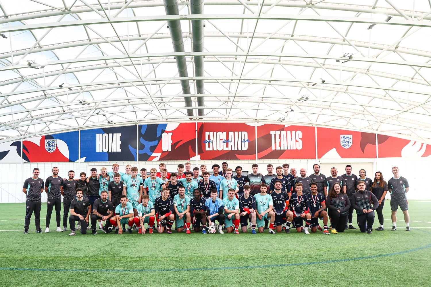 Large group photo of soccer teams and staff lined up on indoor training field with England branding.