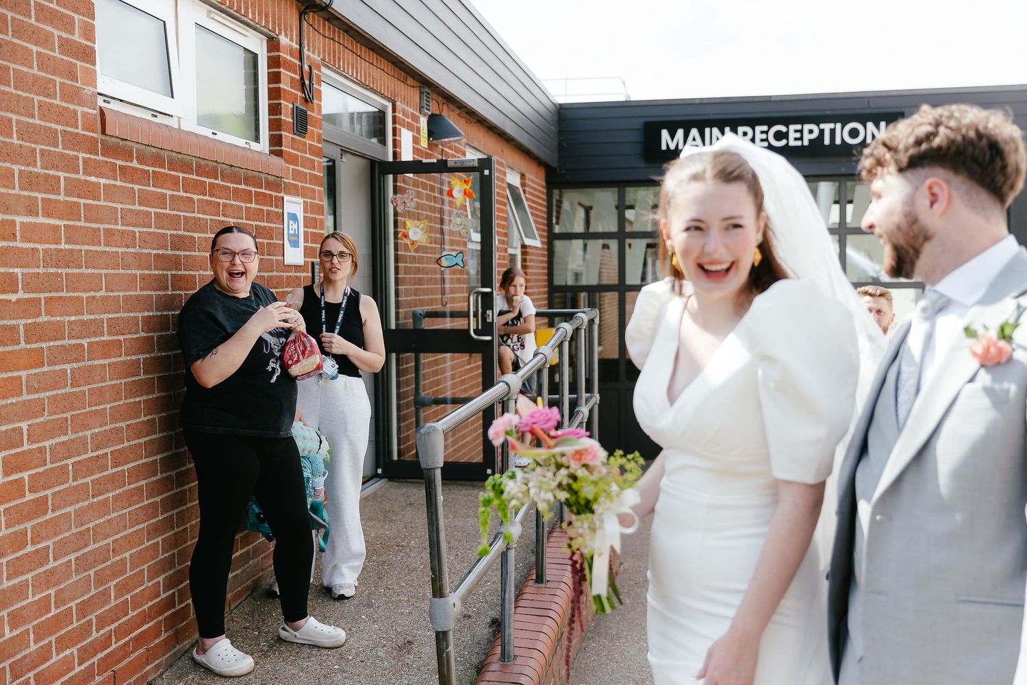 Laughing bride in white dress and veil with groom outside main reception entrance as guests celebrate.