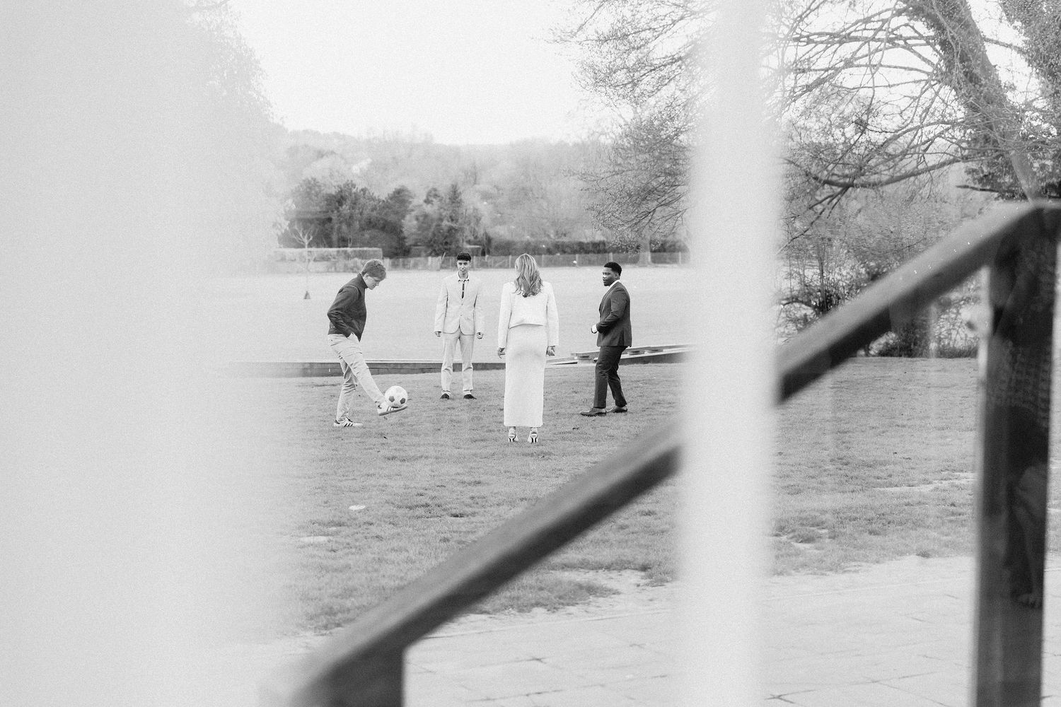 Black and white photo of four people gathered on a lawn near a lake, with bare trees in the background.