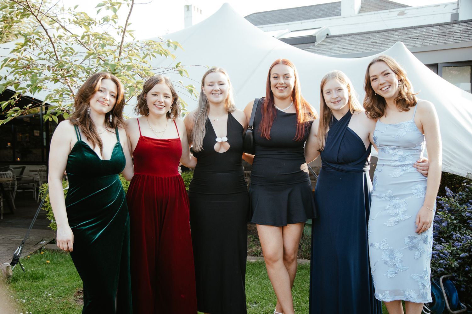 Friends in formal dresses pose together at an outdoor gathering.