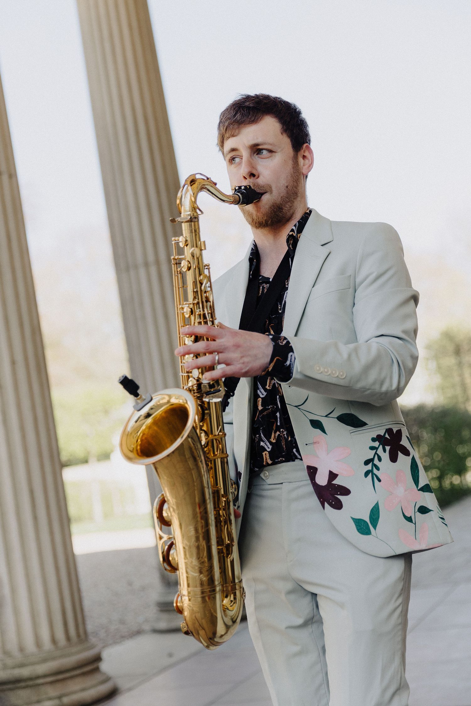 Man in light suit plays saxophone outdoors near white columns, wearing floral-accented jacket.
