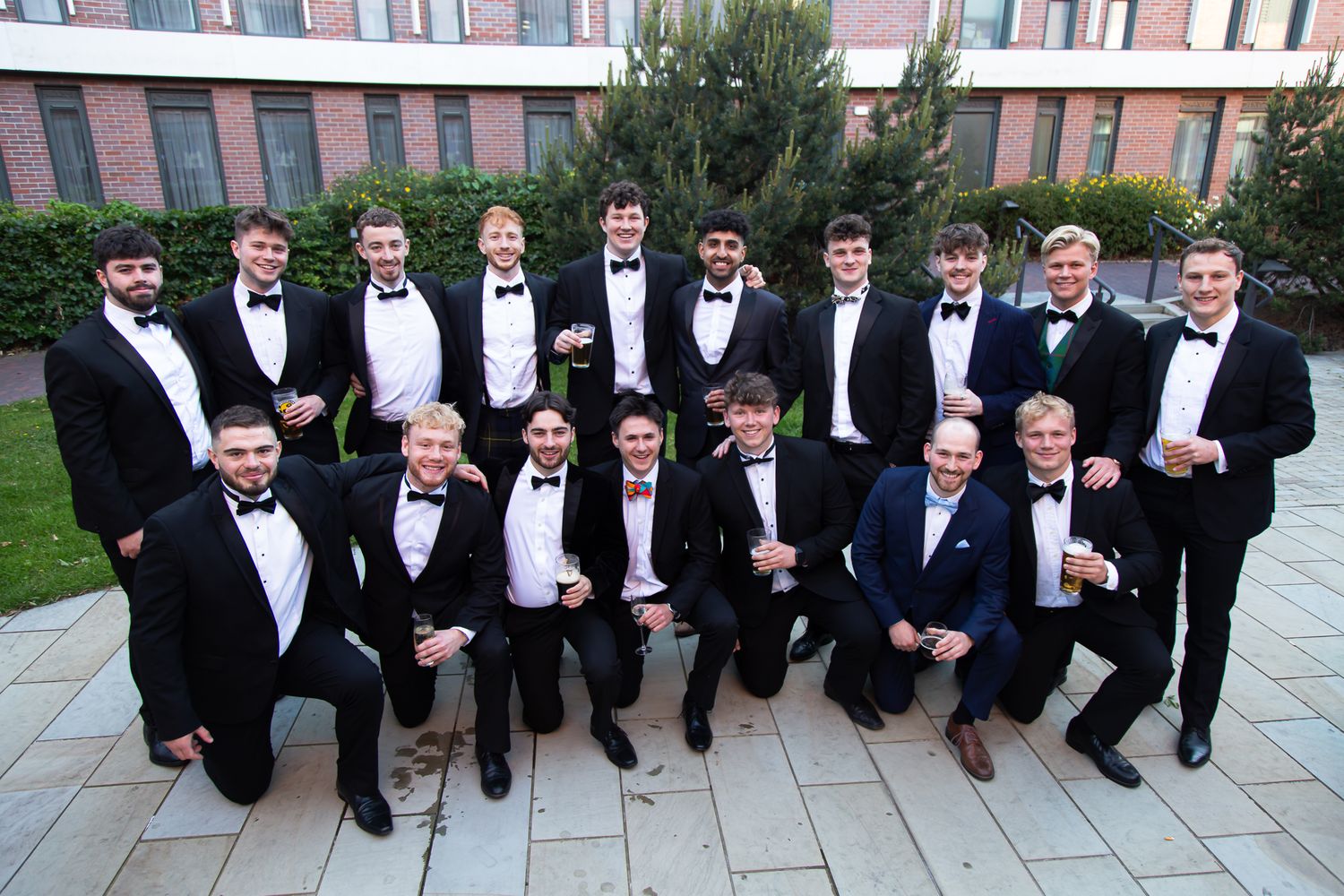 Formal group photo of attendees in tuxedos lined up outdoors.