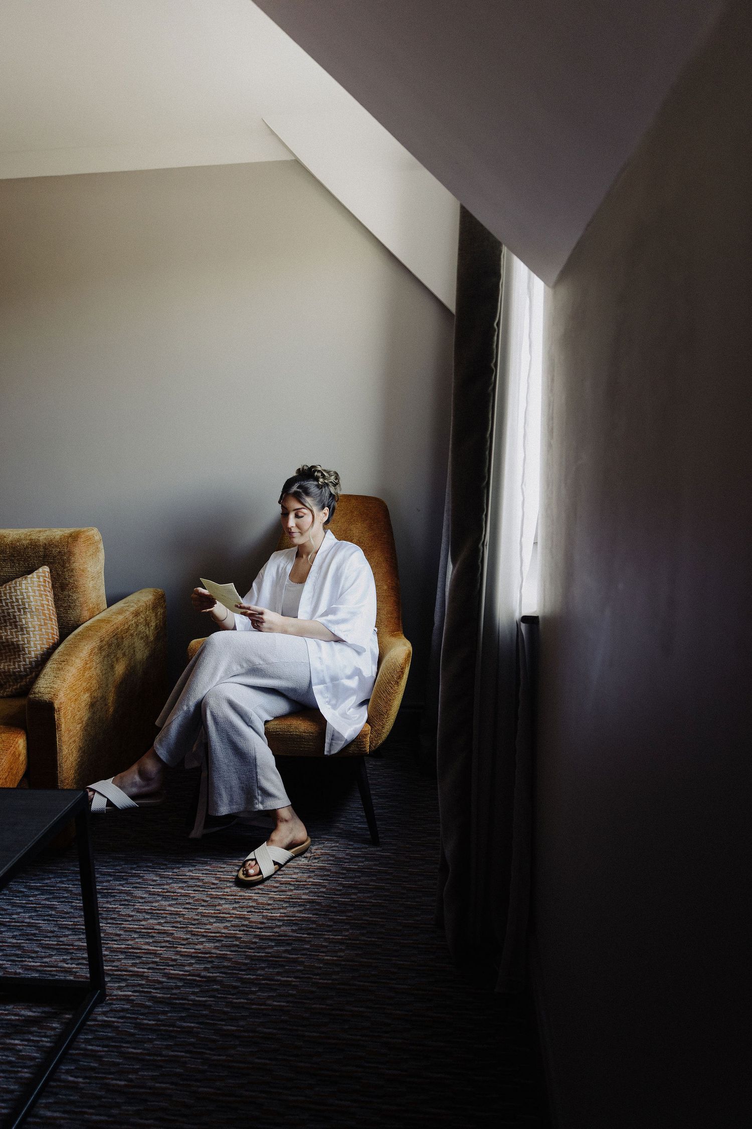 Woman in white robe sits in yellow chair reading near window in modern hotel room.