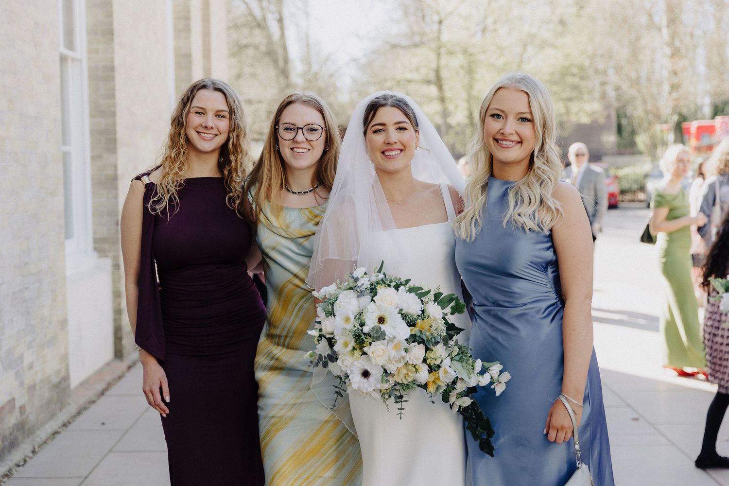 Bride holding white bouquet poses with three bridesmaids in purple and blue dresses outdoors.