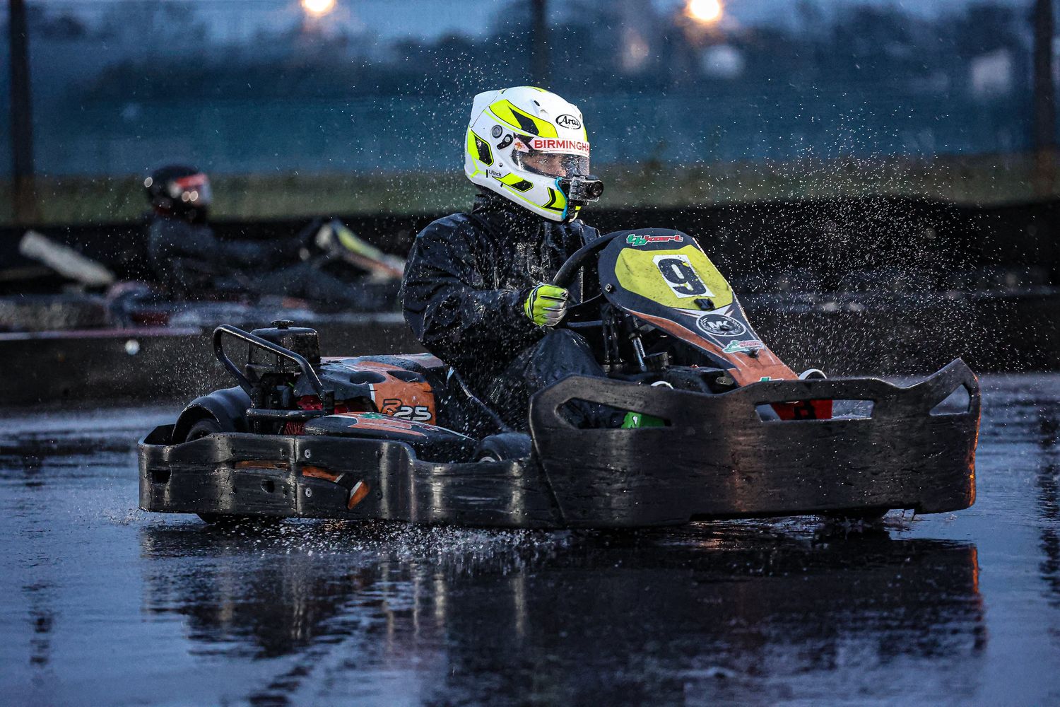 A go-kart racer splashing through wet conditions during a race at dusk.