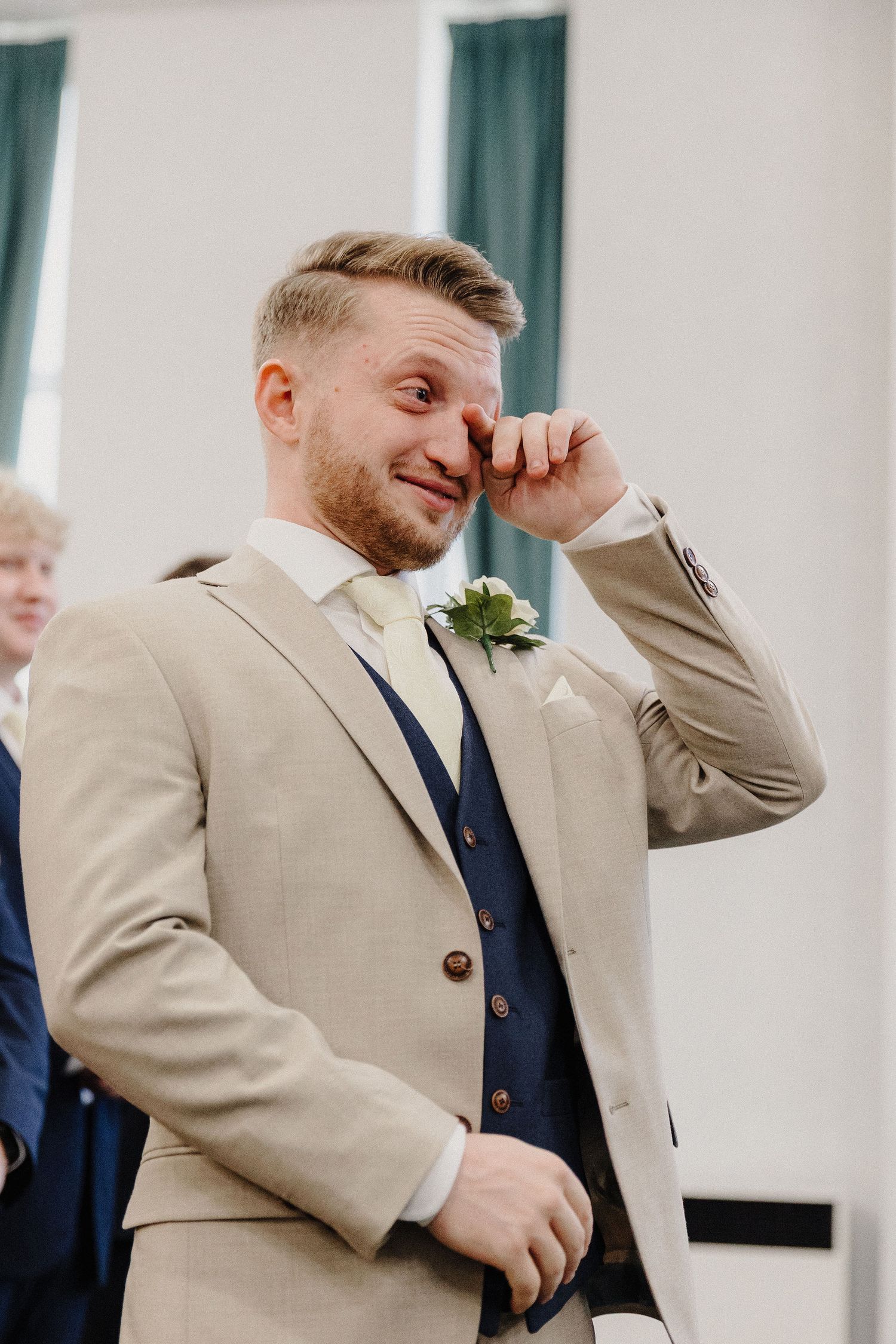 Emotional groom in beige suit wiping tears at wedding ceremony with boutonniere.