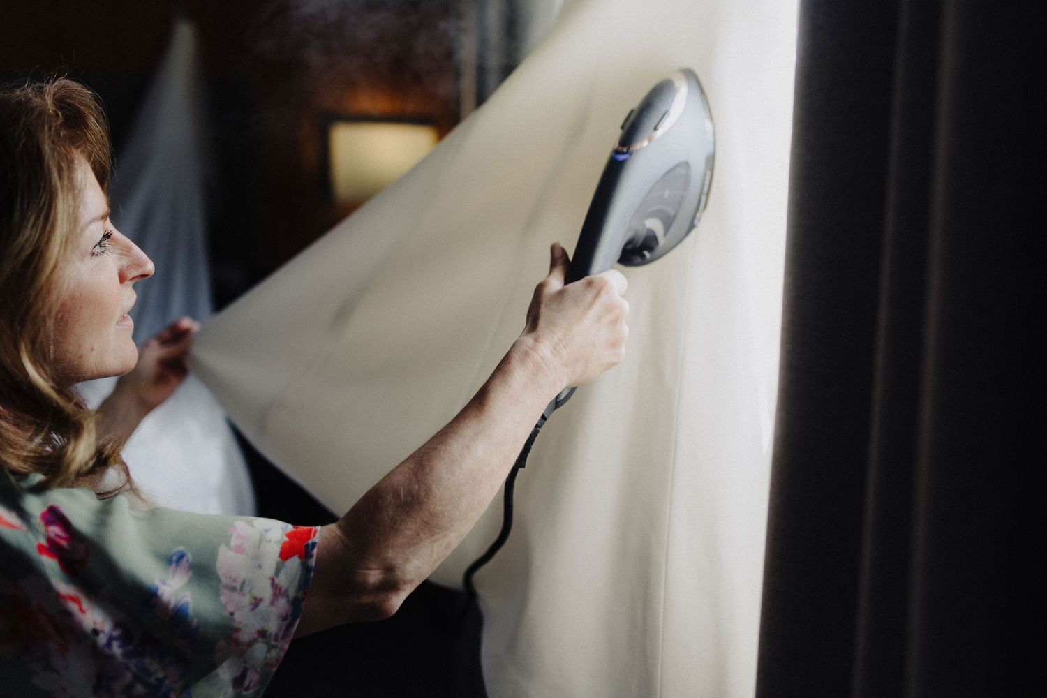 Woman steaming a white curtain or fabric using a handheld garment steamer in a dimly lit room.