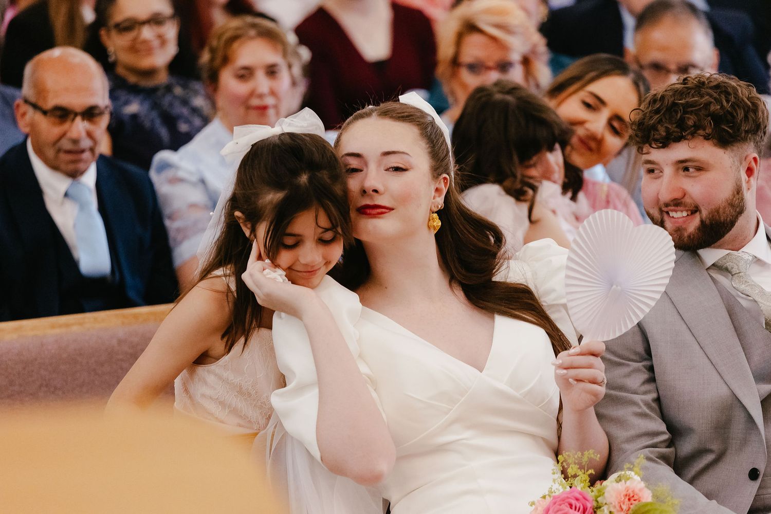 Bride sits with a young girl and groom during wedding ceremony, holding a paper fan and bouquet of flowers.