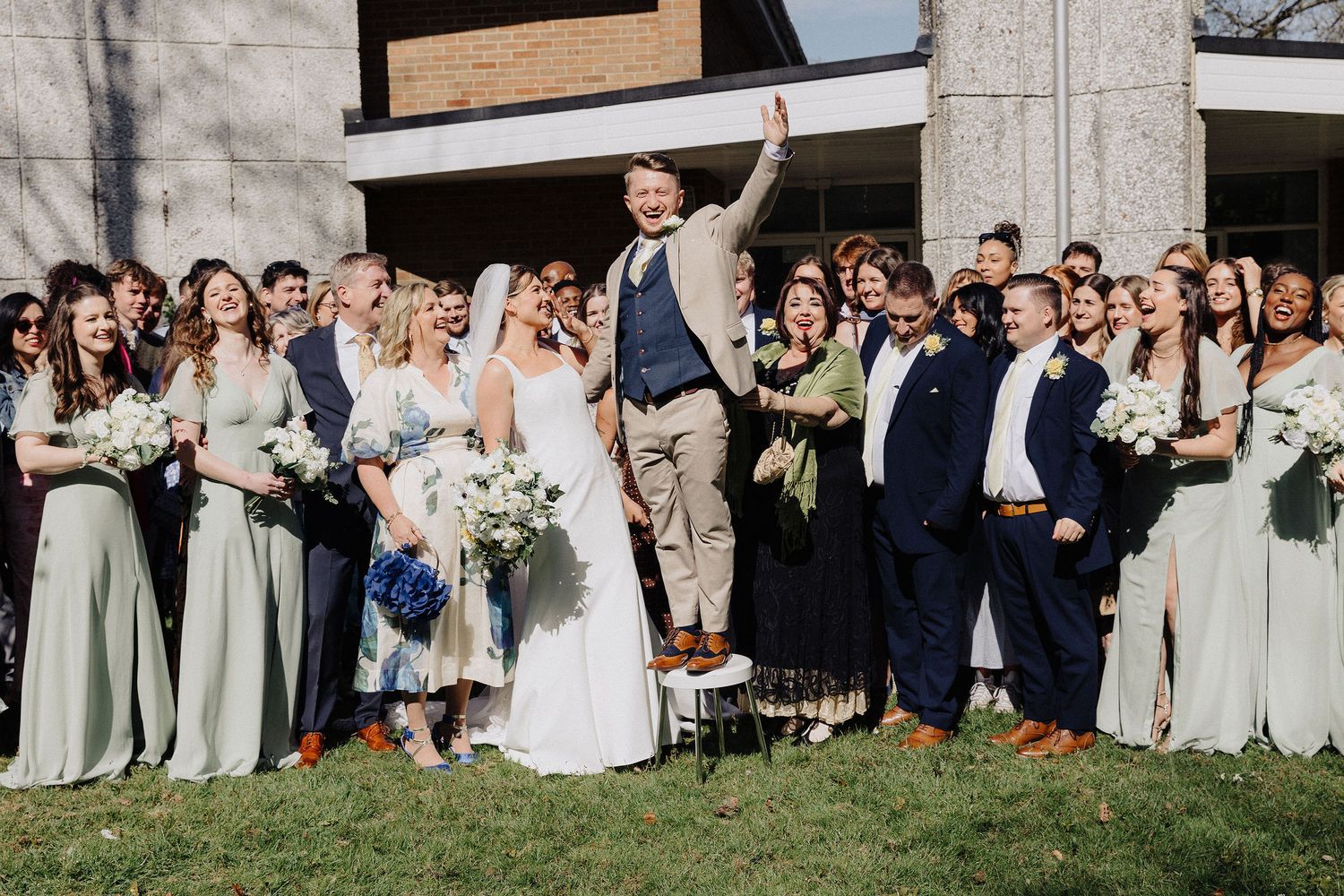 Large wedding party celebrating outdoors with bride and groom, bridesmaids in sage green dresses and groomsmen in navy suits.