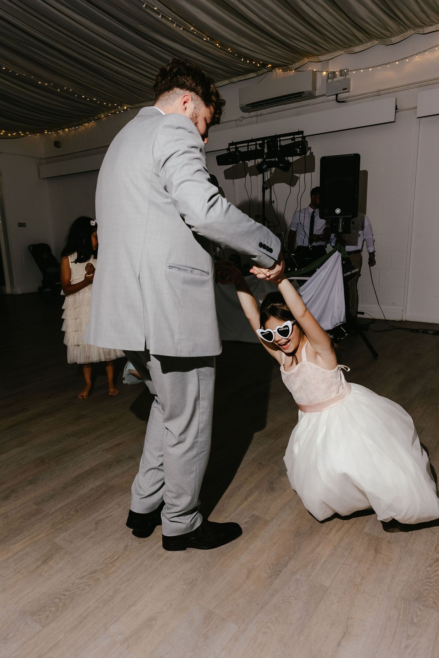 Flower girl in sunglasses dancing with groomsman in gray suit at a wedding reception.