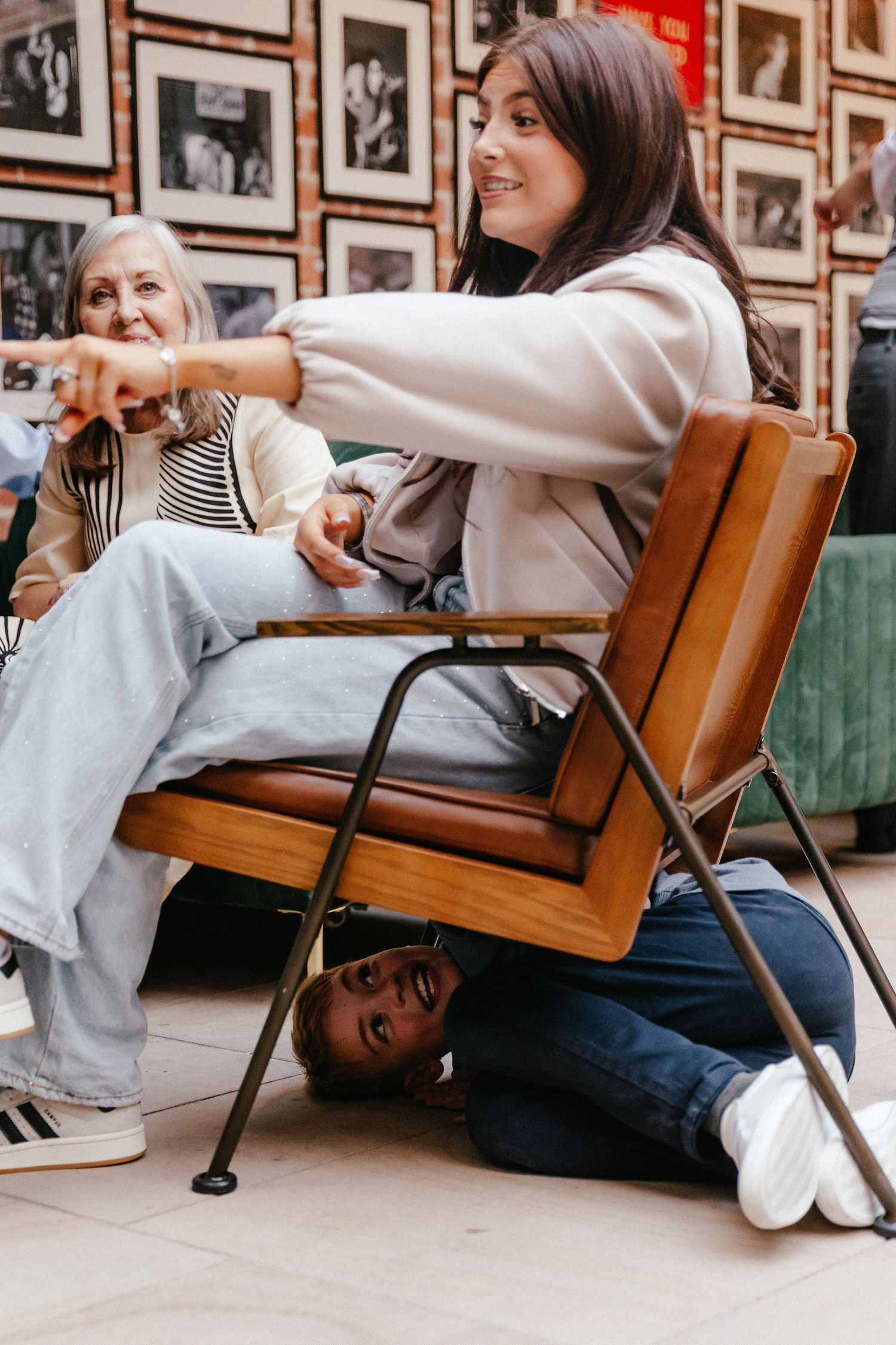 A young woman sits in a leather chair pointing forward while a man lies under it and an older woman sits nearby.