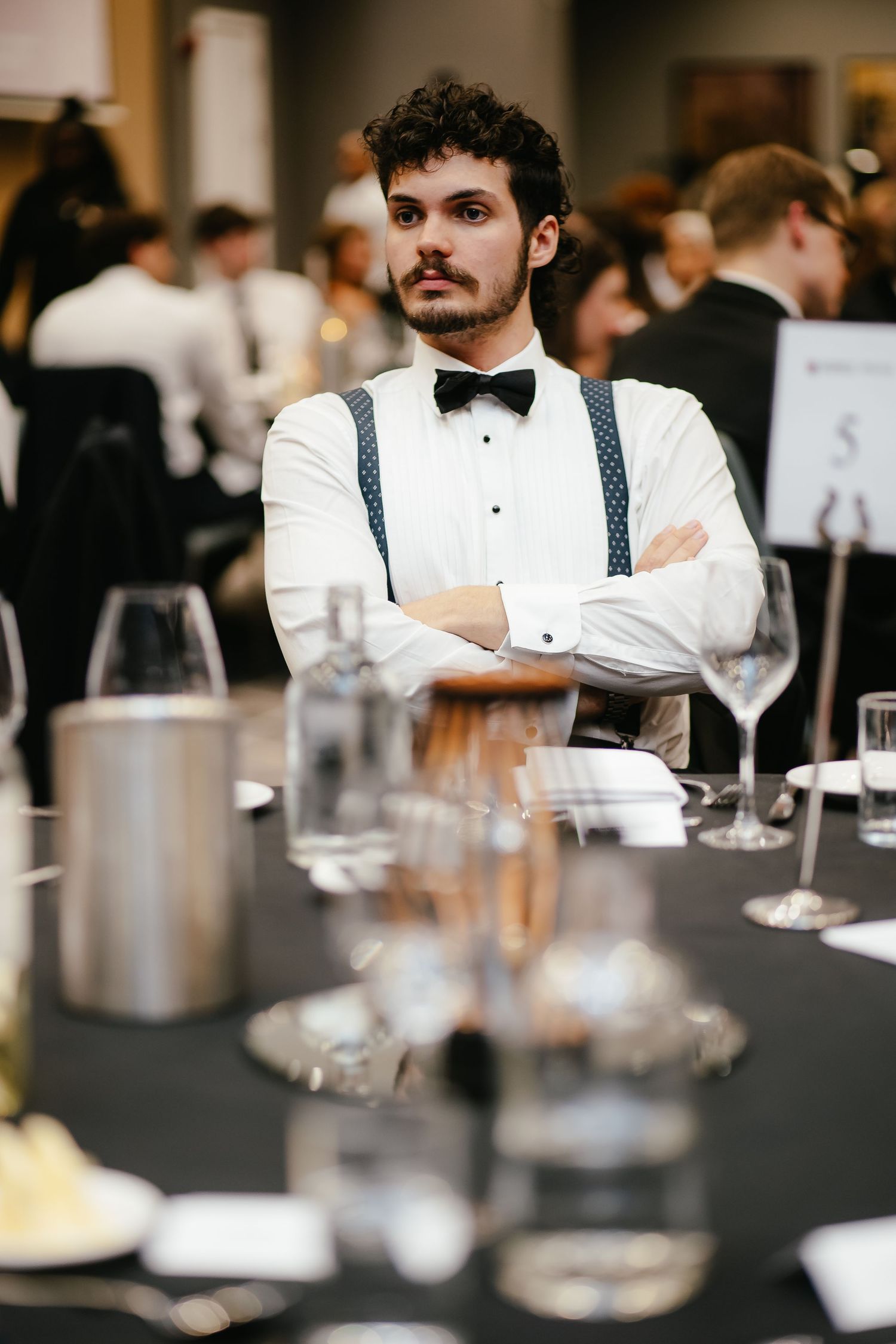 Formal attendee in bow tie and suspenders poses at elegant dinner table setting.