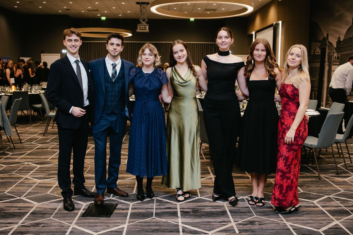 A group of young people dressed in formal attire stand together at an elegant event with geometric carpet patterns.