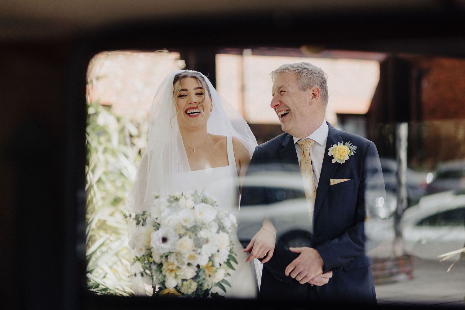 Laughing bride holding white bouquet stands with older man in navy suit, photographed through vintage car window.