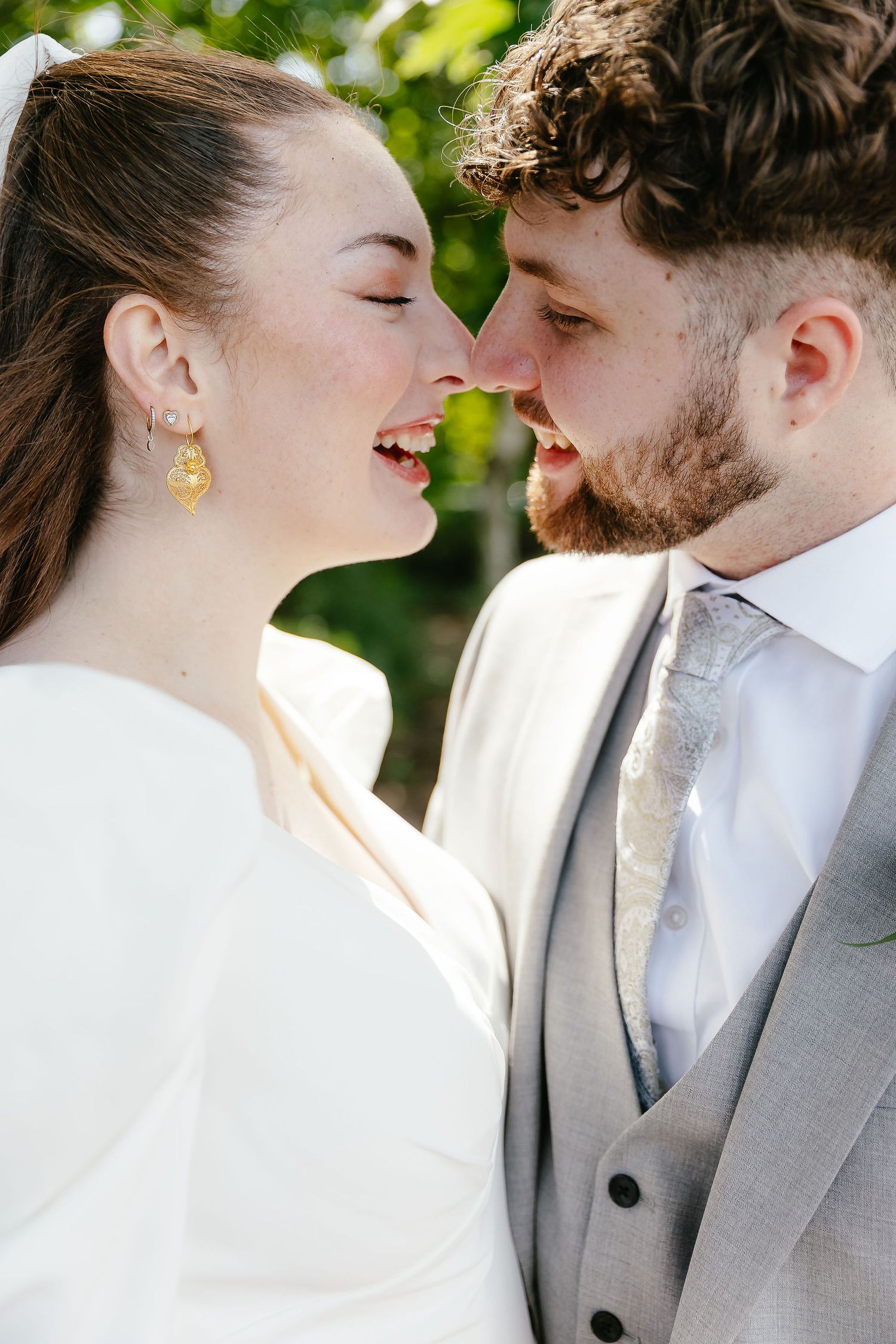 Bride and groom share a joyful nose-to-nose moment outdoors, bride wearing gold earrings and white dress.