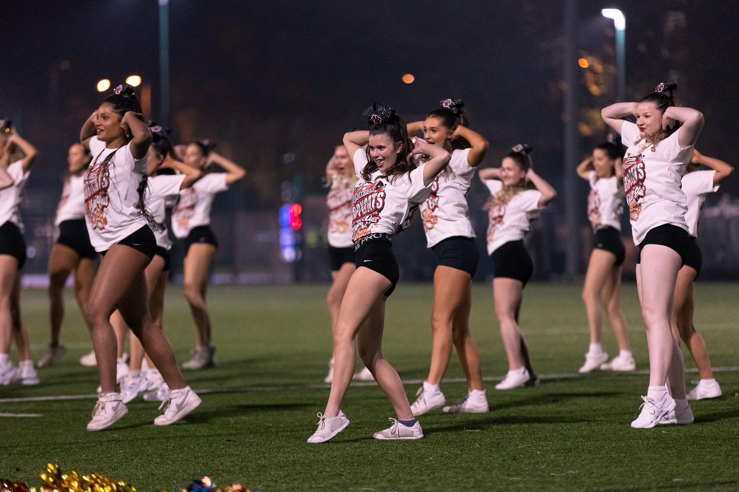 Cheerleaders in white uniforms perform synchronized dance moves on a grassy field at night.