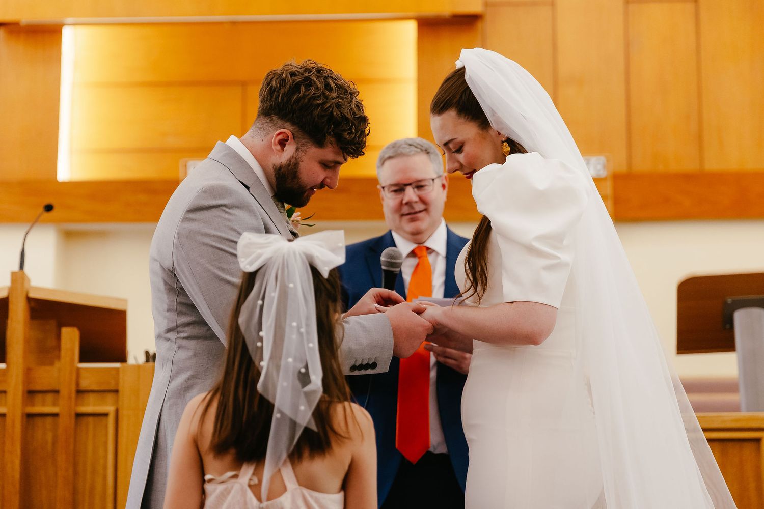 Bride and groom exchange rings at altar with officiant and flower girl during an elegant church wedding ceremony.