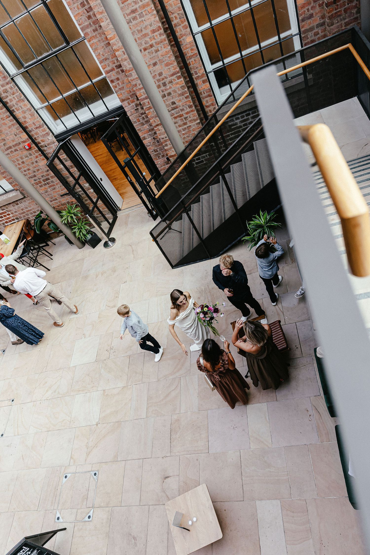 Aerial view of wedding guests gathering in a modern brick atrium with staircases, as bride holds bouquet.
