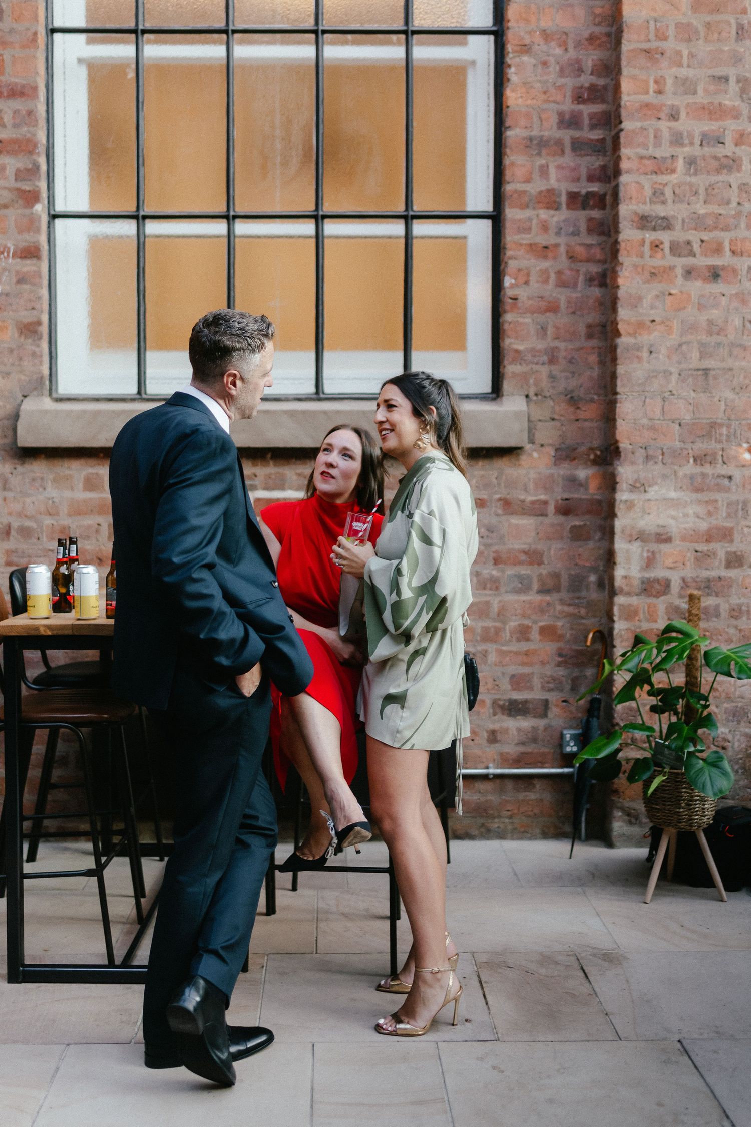 Three elegantly dressed people socializing at an upscale indoor venue with exposed brick walls.