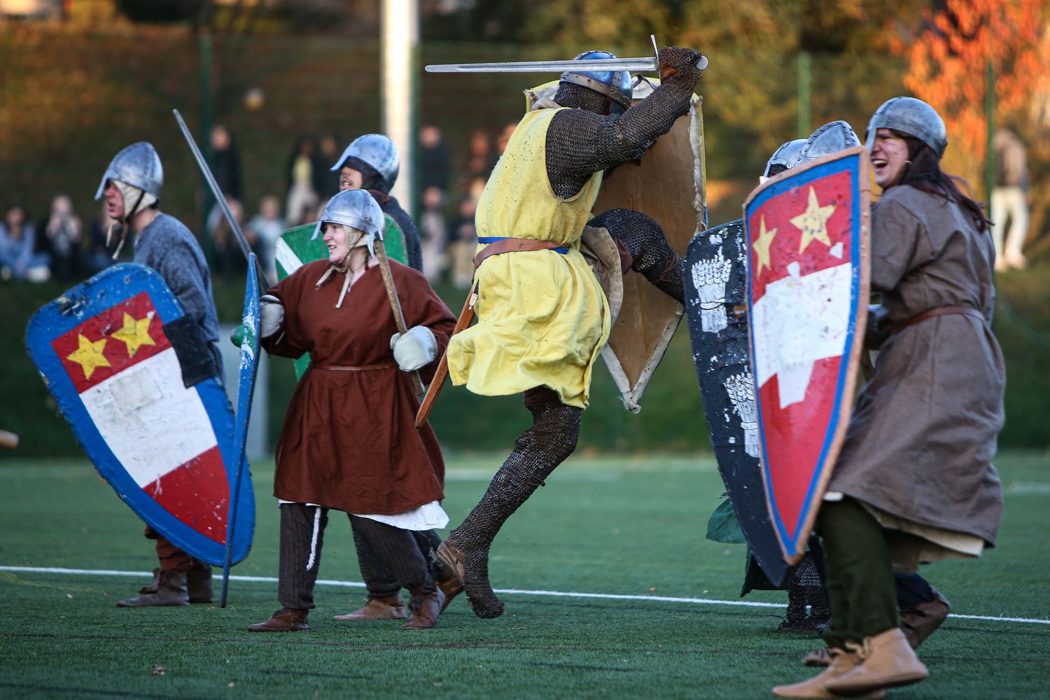 Medieval costumed performers with shields and armor engage in combat reenactment during an outdoor event.