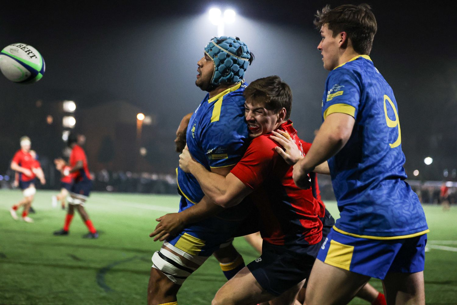 Rugby players from opposing teams compete for possession of the ball during a night match under stadium lights.