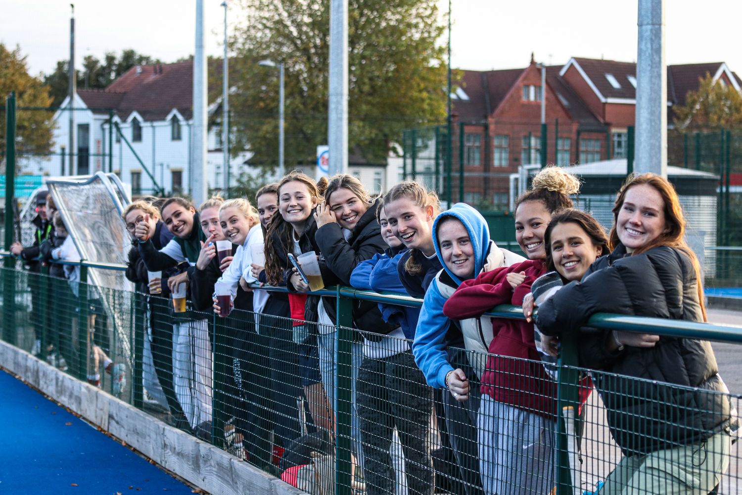 A group of spectators line up along a fence to watch a sporting event on a sunny day.