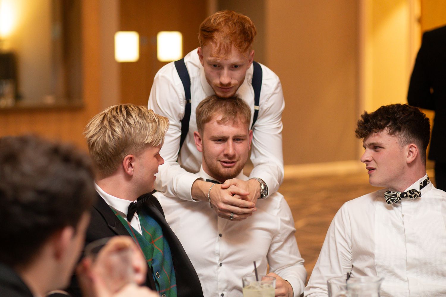 Group of formal-dressed attendees laughing and embracing at an indoor celebration event.