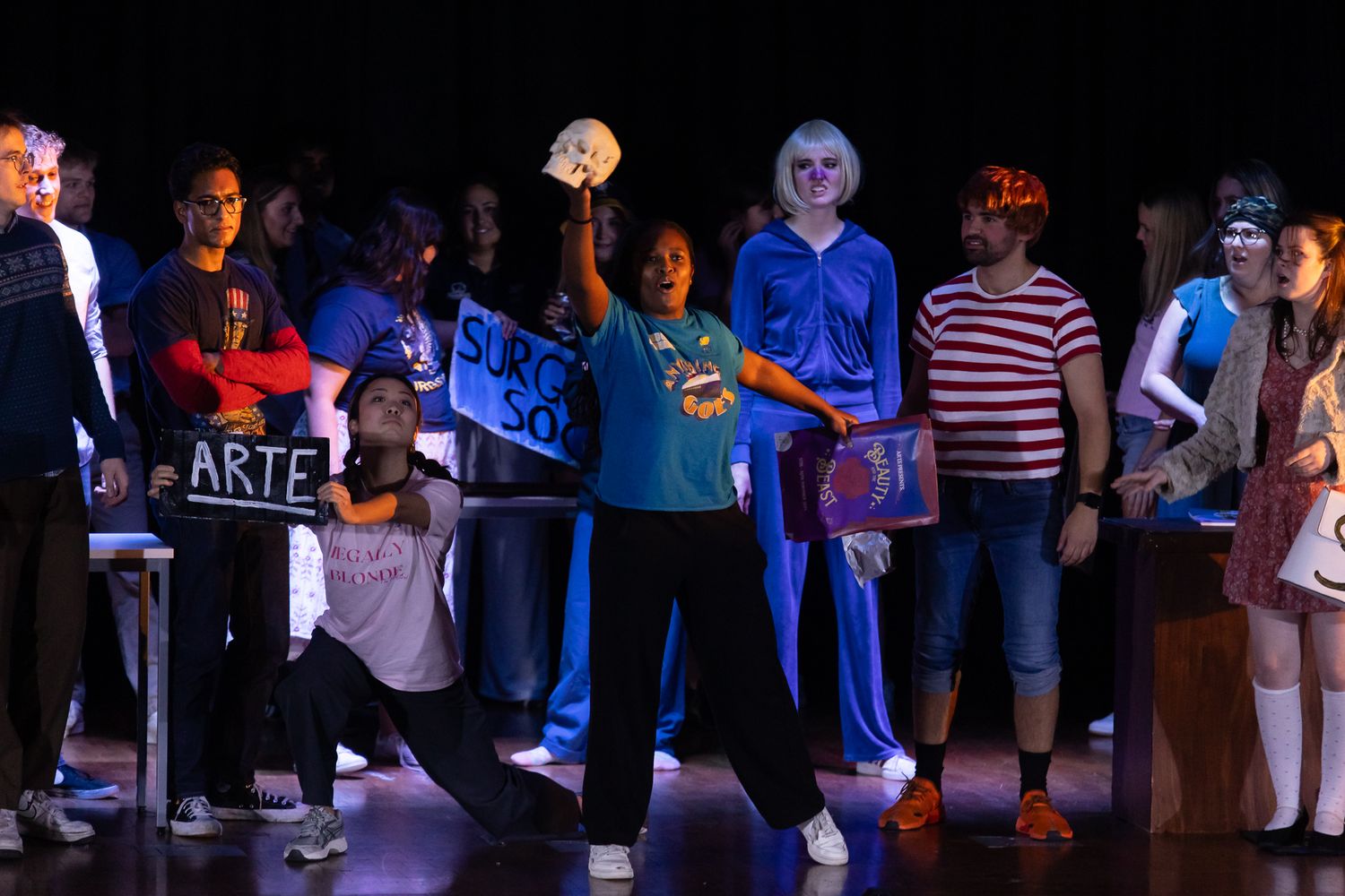 Theater performers in various costumes under dramatic blue stage lighting.