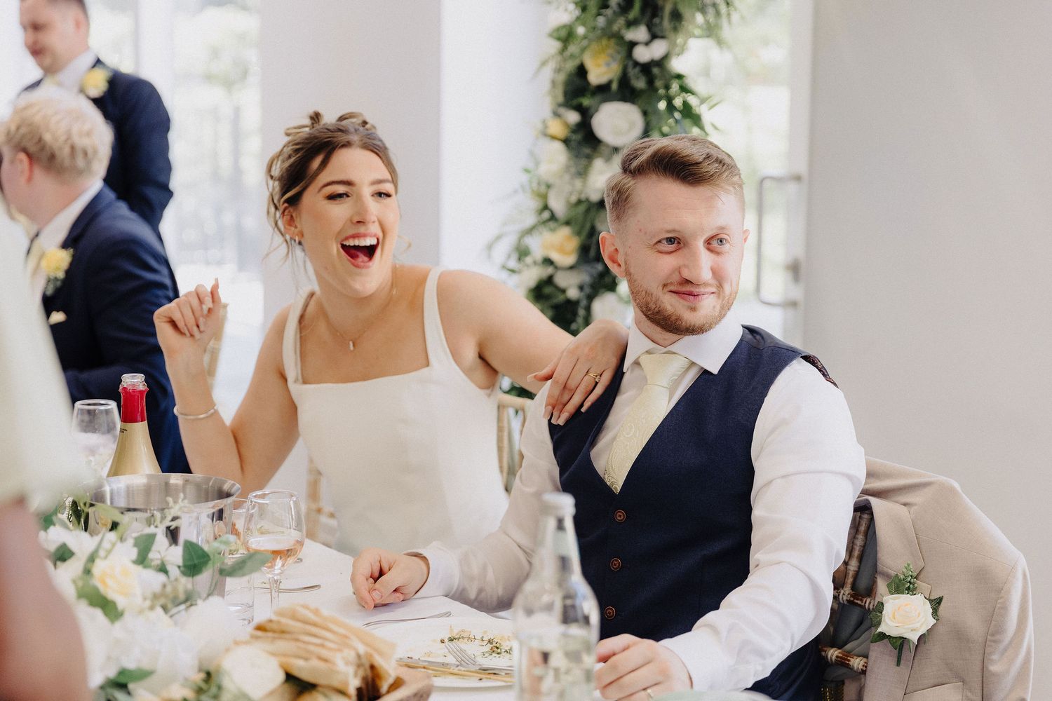 Bride and groom laughing joyfully at wedding reception dinner table, decorated with flowers and candles.