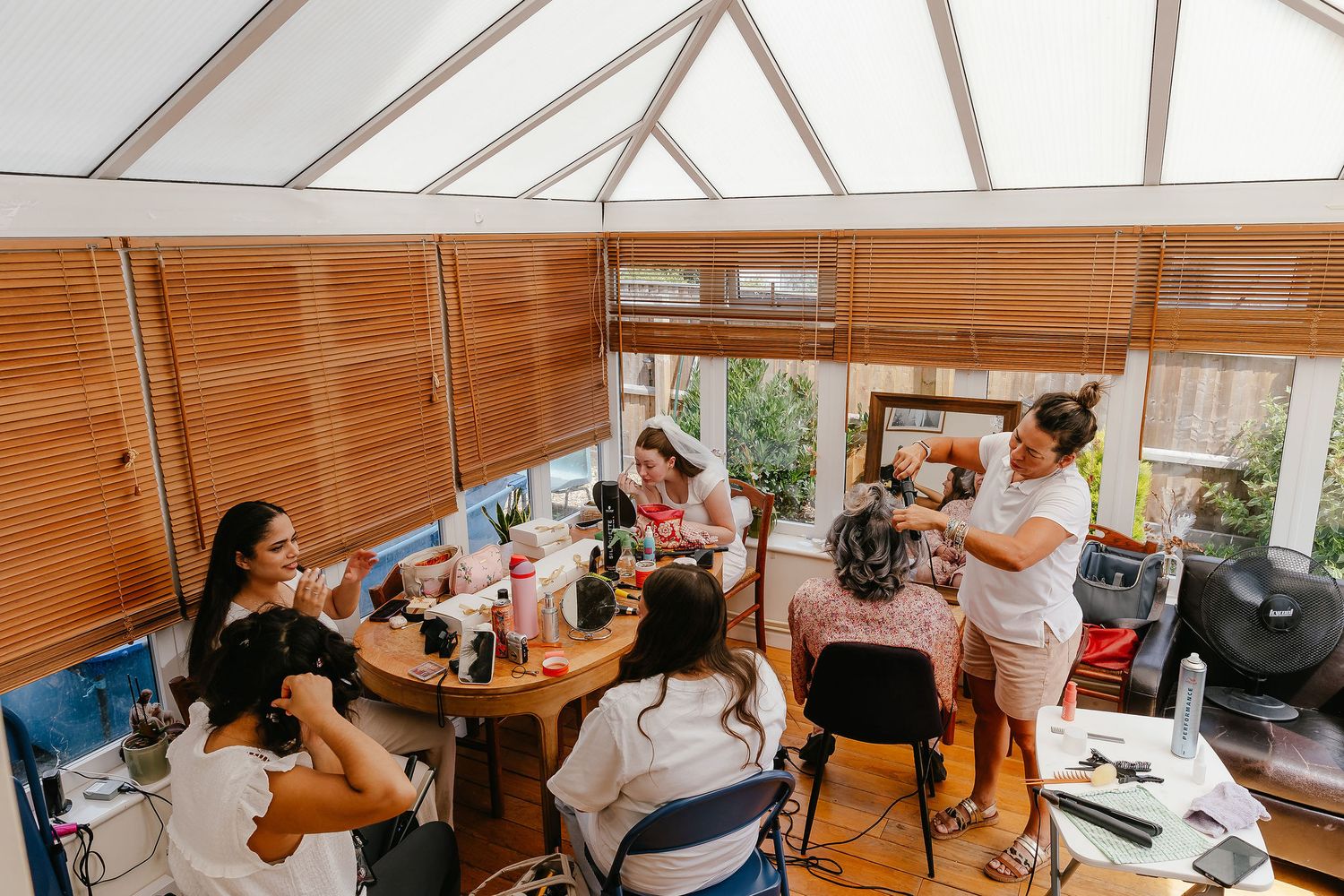 Women gathered around a table in a sunroom getting hair and makeup done for a special occasion.