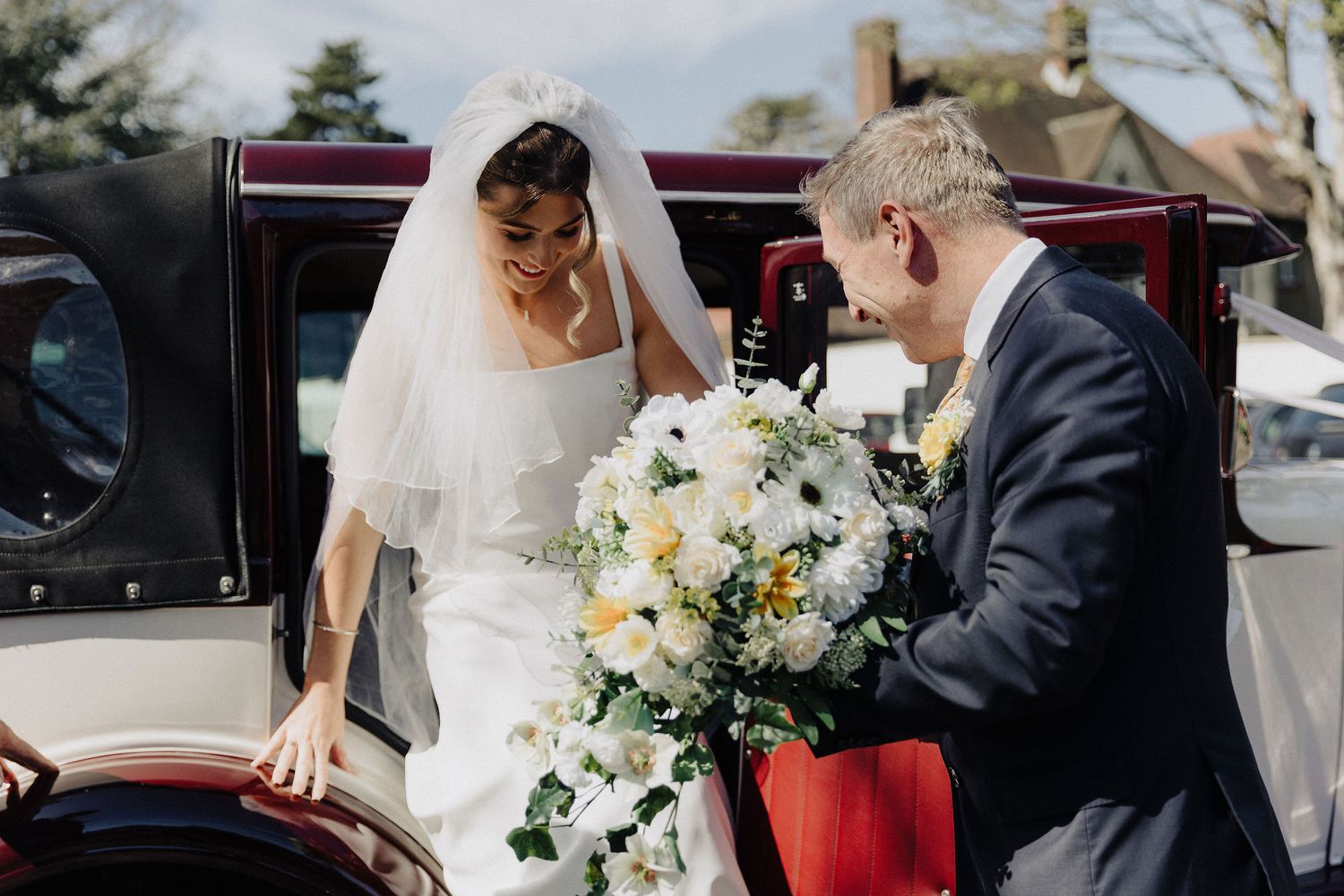 Bride in white gown receiving bouquet from groom beside vintage wedding car.