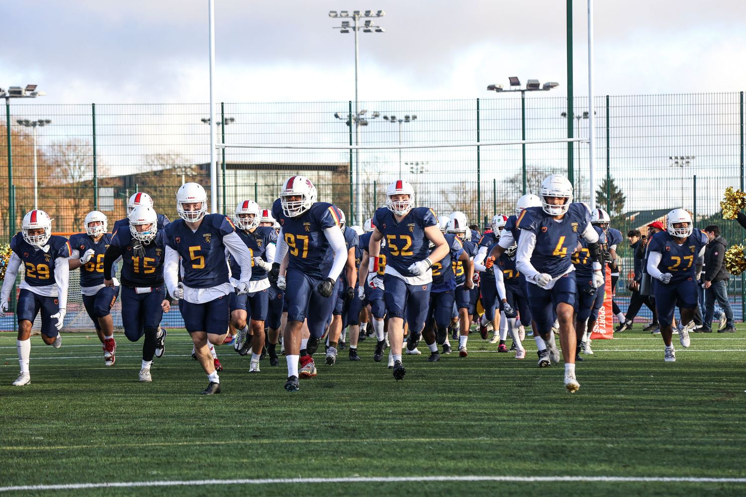 Football players running drills on an outdoor turf field during team practice.