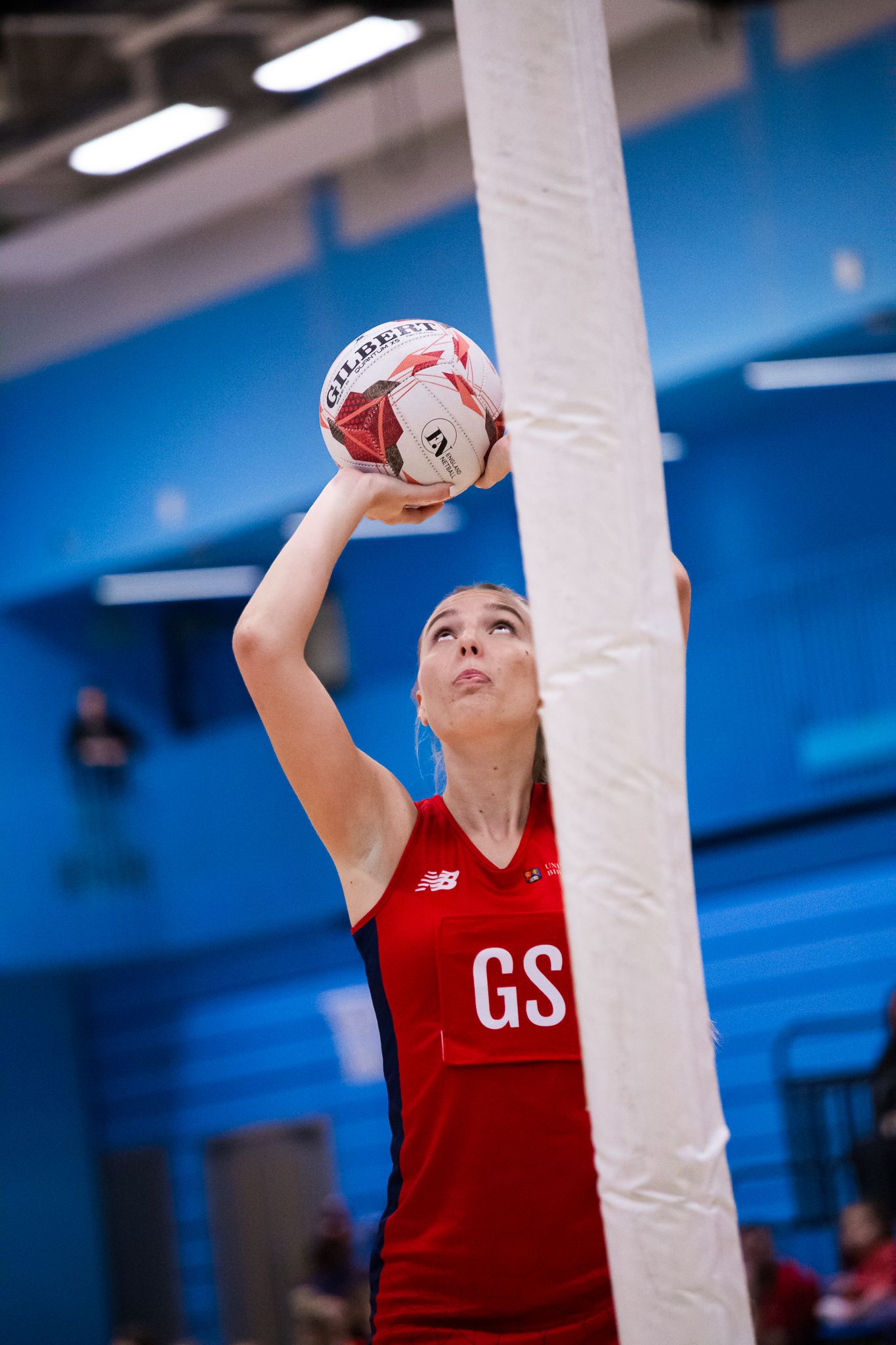 Netball player in red uniform practicing shooting drills in an indoor court.