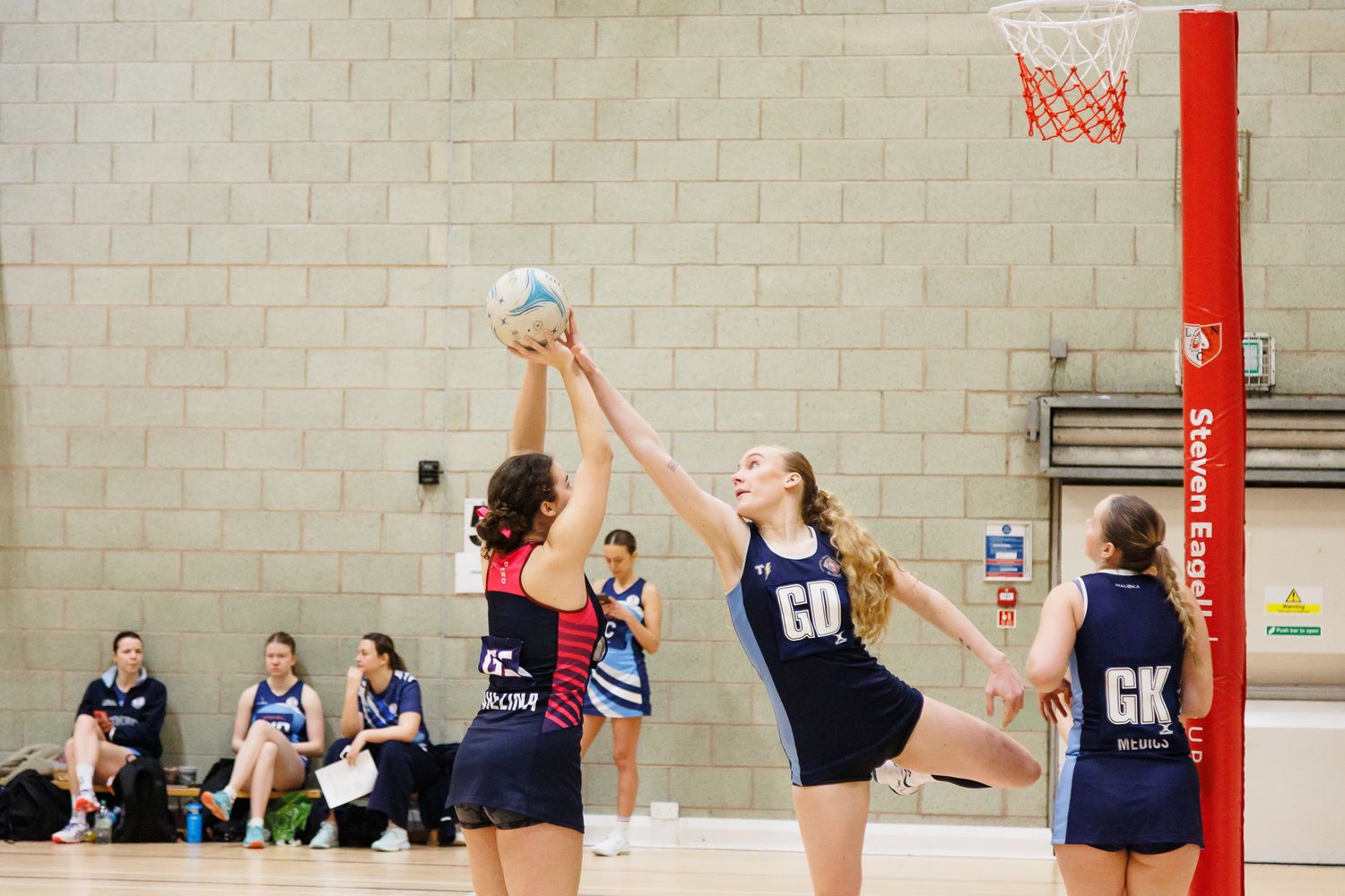 Indoor netball court with players practicing near a red goal post.