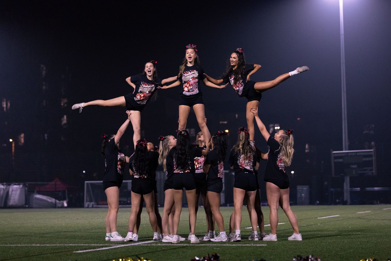 Cheerleaders perform a dramatic nighttime pyramid stunt formation on an illuminated sports field under bright stadium lights.