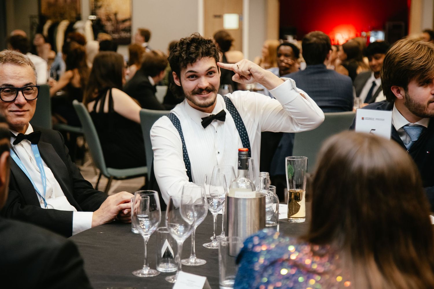 Guests in formal attire enjoying conversation at a dinner table with wine glasses.