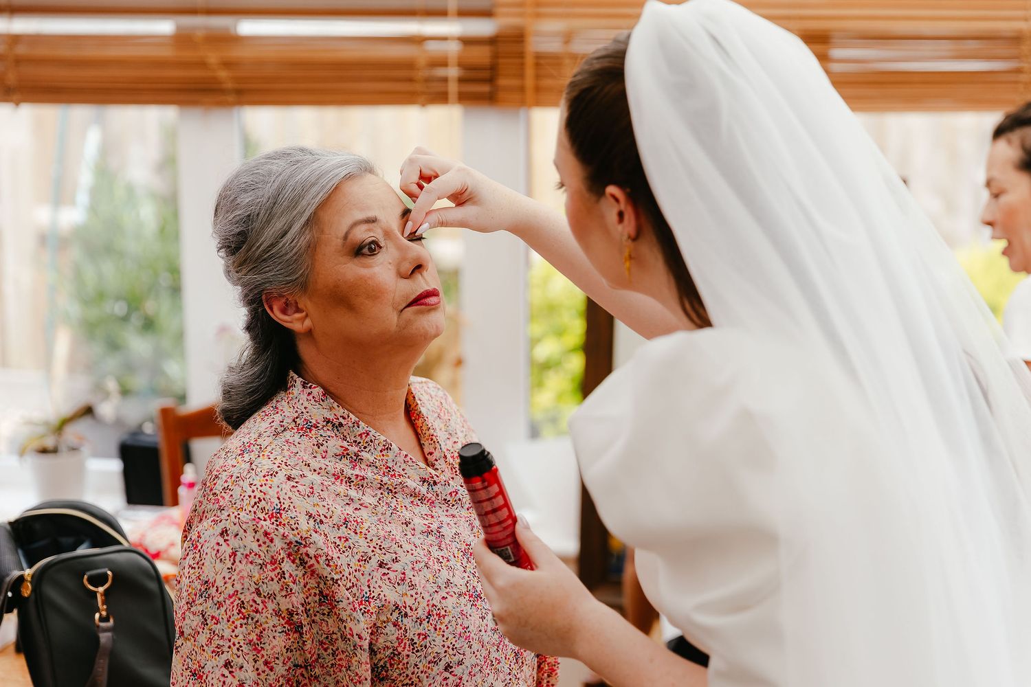 Bride in white veil applying makeup to older woman in floral dress during wedding preparation.