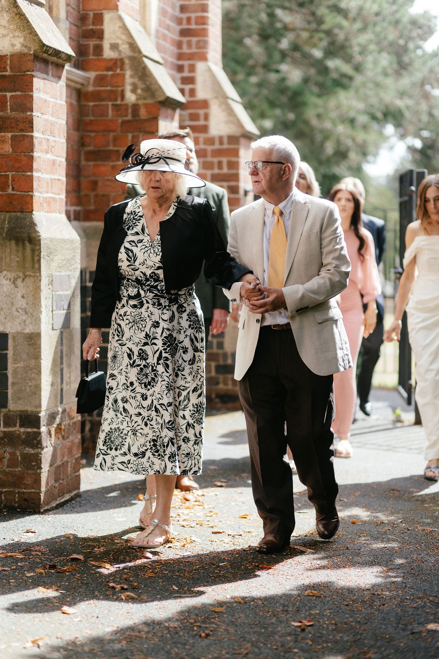Elderly couple in formal attire walking together at a wedding, woman in floral dress and hat, man in beige suit.