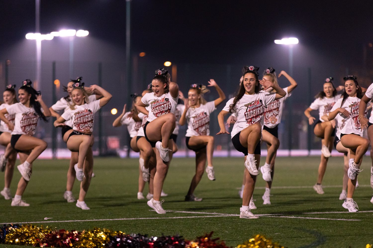 A group of cheerleaders perform a synchronized high kick line routine on a field at night under bright stadium lights.