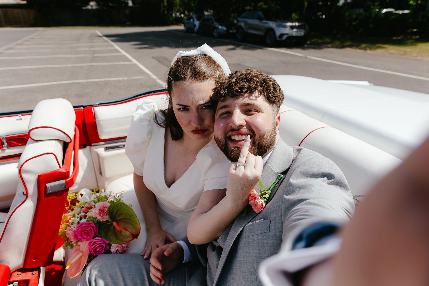 Bride and groom laughing in a vintage red and white convertible car on their wedding day.