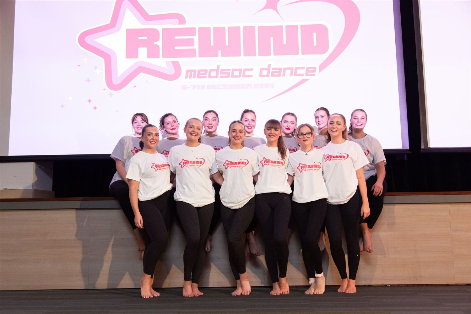 Dance performers posing on stage with pink branded backdrop for group photo.