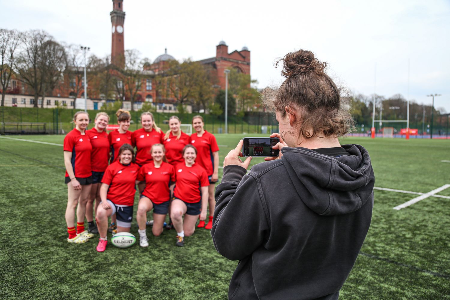 Team gathering on athletic field with brick buildings visible in background.