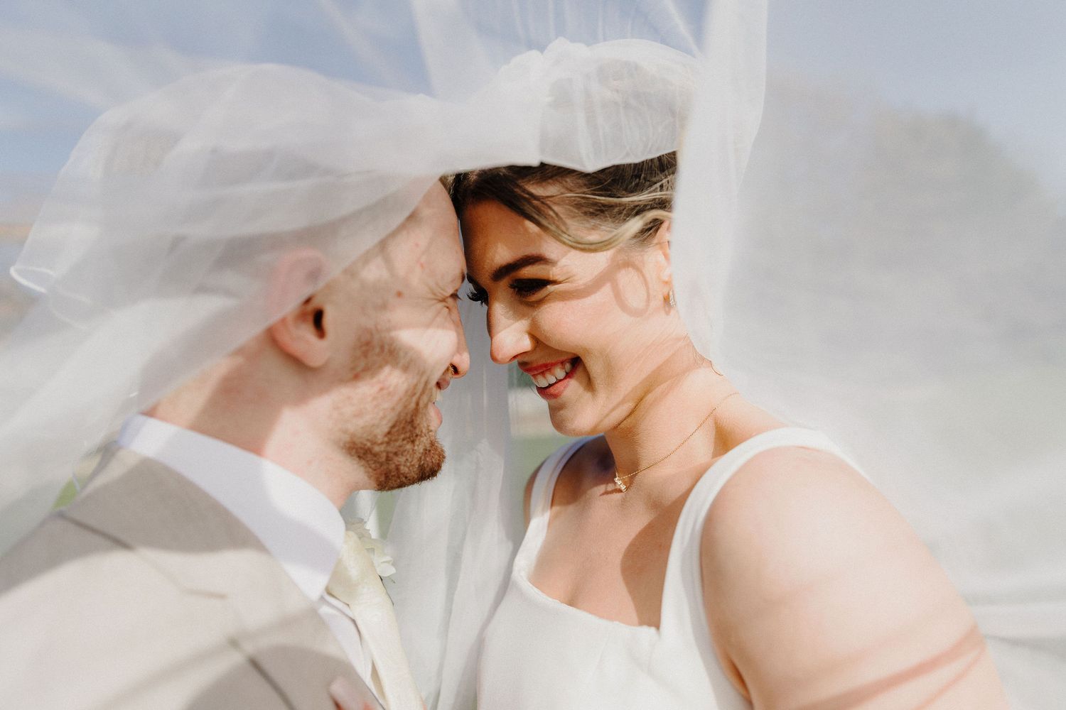 Bride and groom share a romantic moment under a flowing white veil, smiling and touching foreheads on their wedding day.