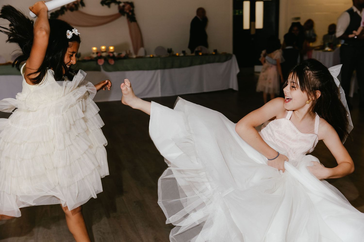 Two girls in white dresses dancing joyfully at a wedding reception on a dance floor.