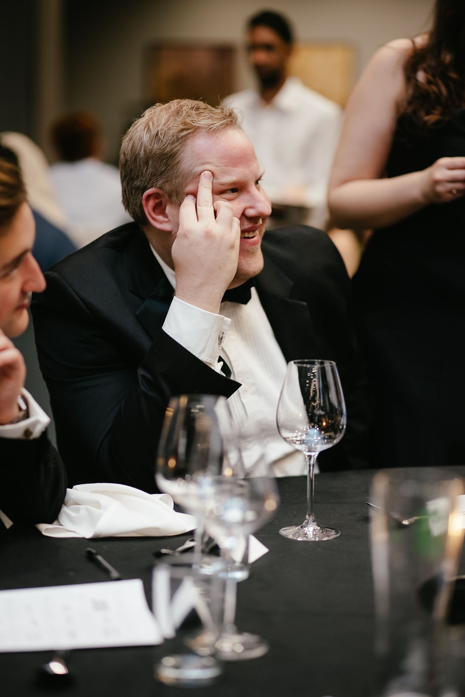 People in formal attire seated at a dark table with wine glasses during an event.