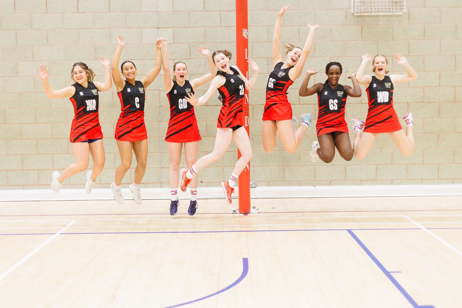 Netball players in red uniforms celebrating together on an indoor court.