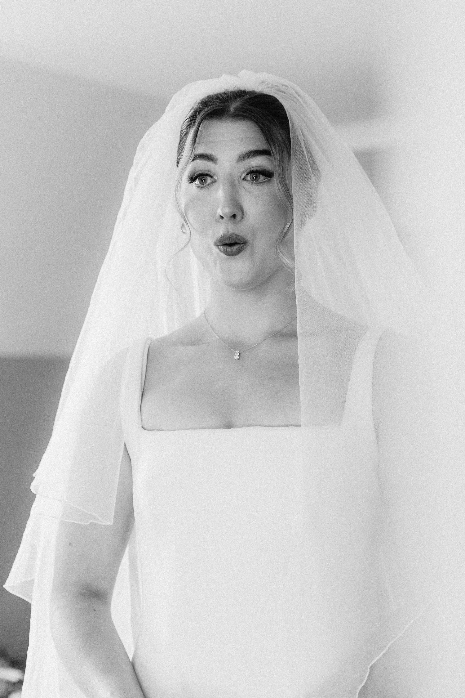 Black and white portrait of bride in square-neck gown and veil making surprised expression indoors.