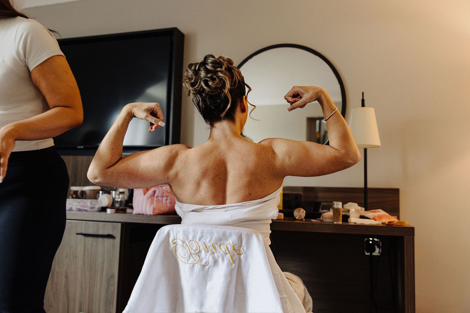 Bride in white wedding dress flexing biceps muscles showing her strong back while getting ready in hotel room.