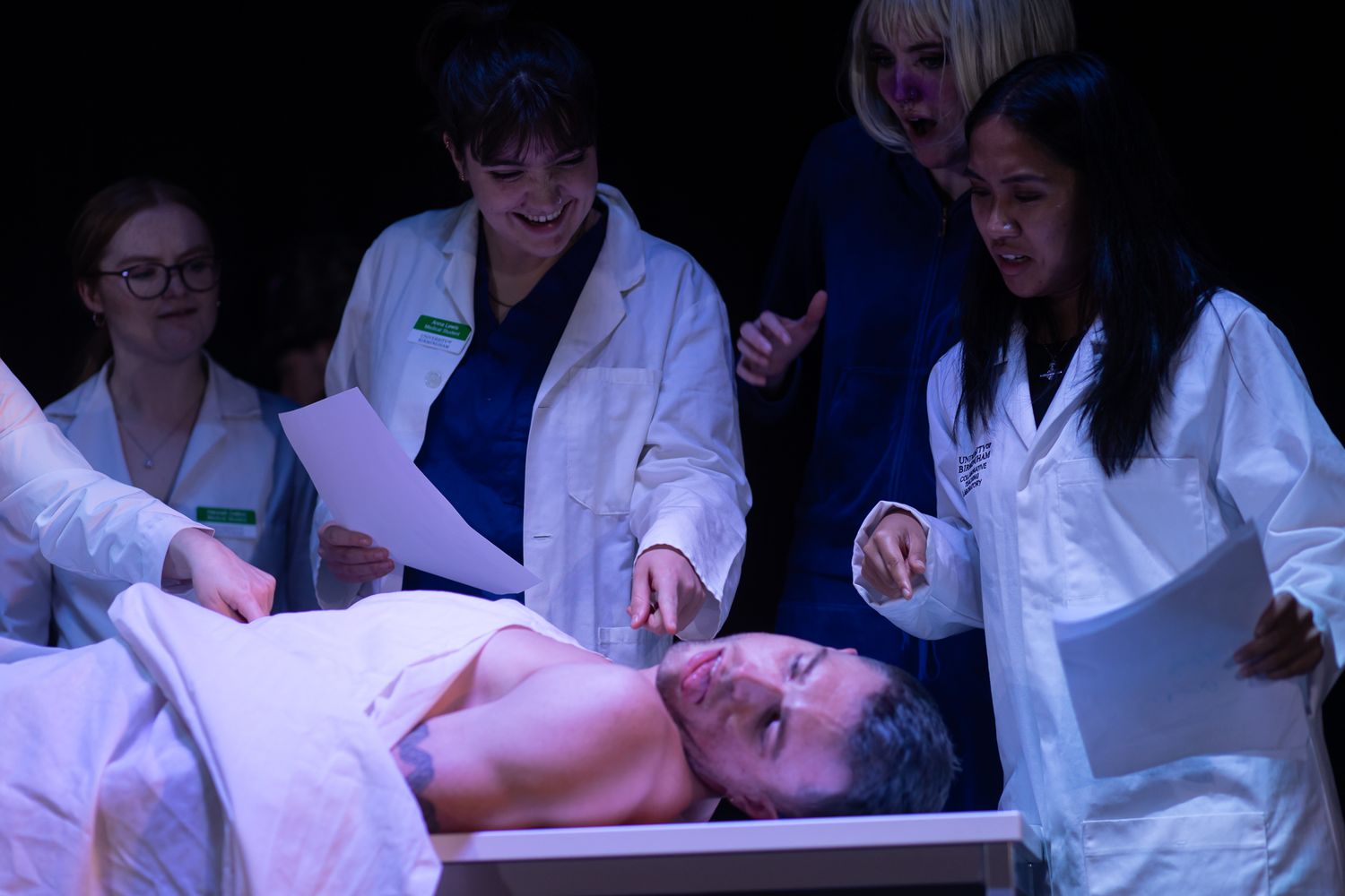 Group of performers in white lab coats interacting on a dimly lit stage.