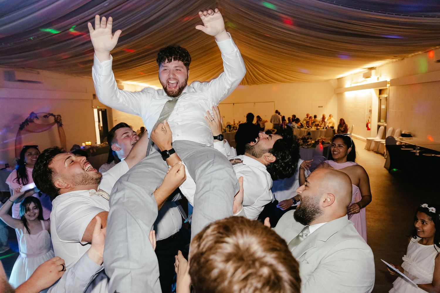 Groom lifted by groomsmen during lively wedding reception dance floor celebration with guests in background.